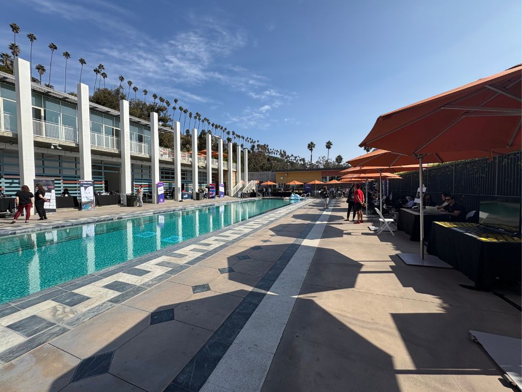 A sunny day at an outdoor poolside area with modern buildings, tall palm trees, people standing by the pool, and orange umbrellas shading tables along the deck.