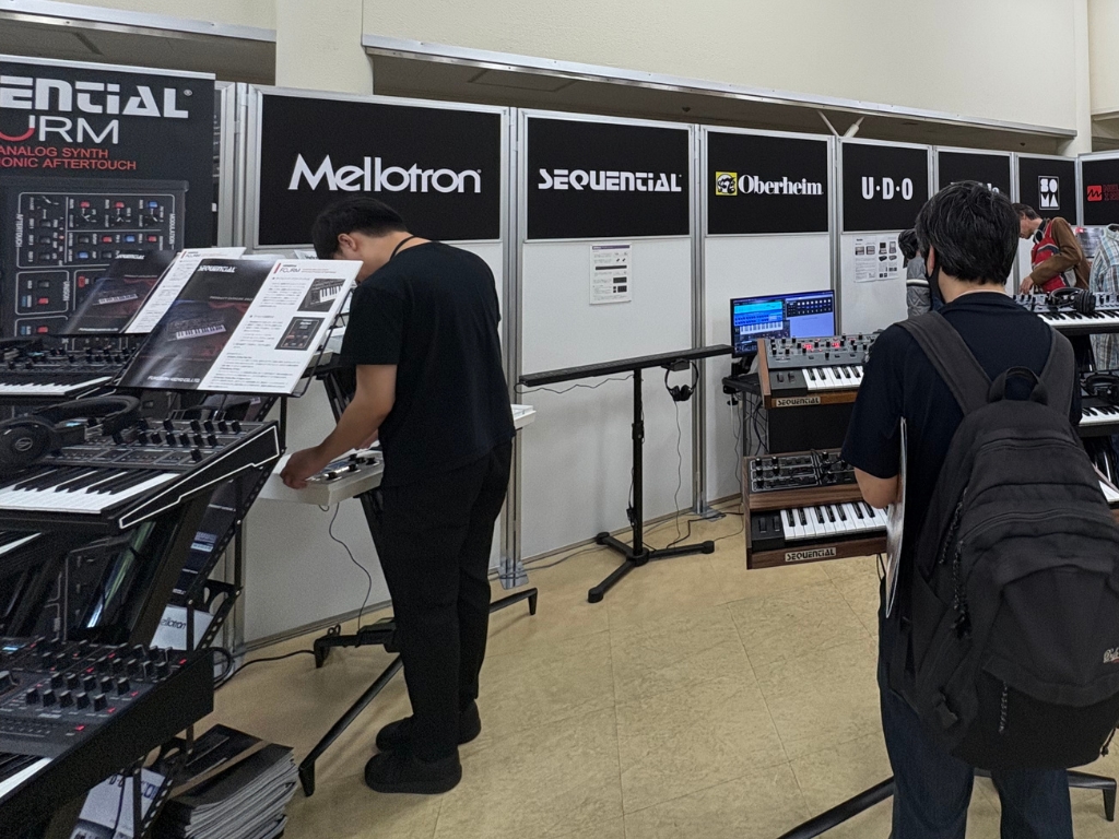 Two people in a music store examine and play synthesizers displayed on stands. Brand signs like Mellotron, Sequential, Oberheim, and UDO are visible on the wall behind them.
