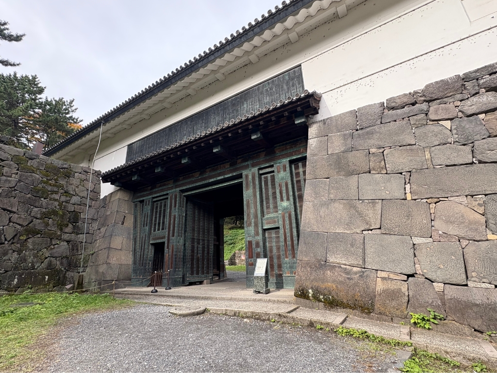 A large traditional Japanese wooden gate with metal details set in tall stone walls, surrounded by greenery and a gravel path, under a cloudy sky.