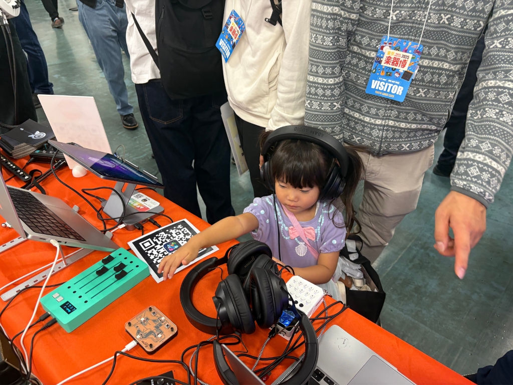 A young girl wearing headphones touches music equipment on a table covered with audio devices and laptops, surrounded by adults at an indoor event.