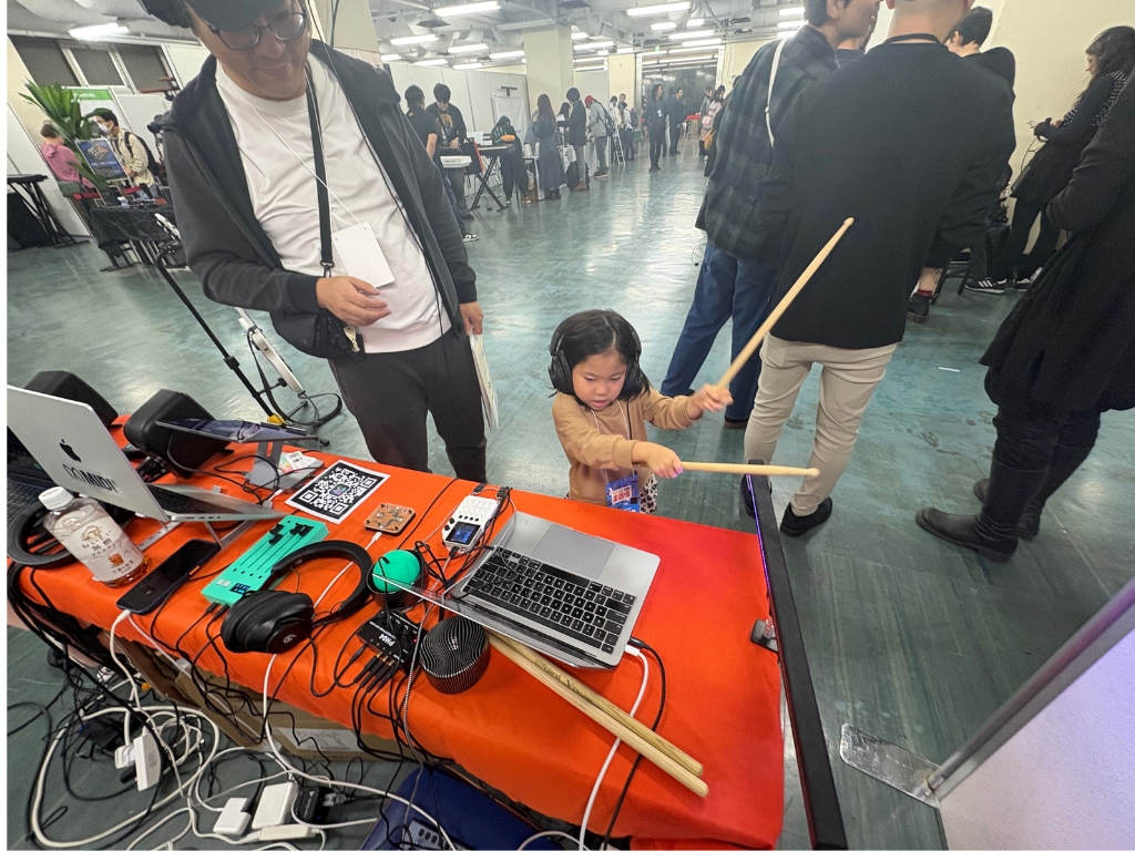 A young child wearing headphones enthusiastically plays with drumsticks near a table with a laptop, cables, and audio equipment. An adult stands nearby. People are gathered in the background of a large indoor space.