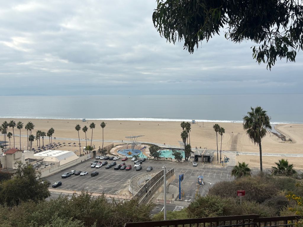 A view of a wide sandy beach with palm trees, a parking lot, a pool area, and a few buildings, under a cloudy sky. The calm ocean stretches into the distance.
