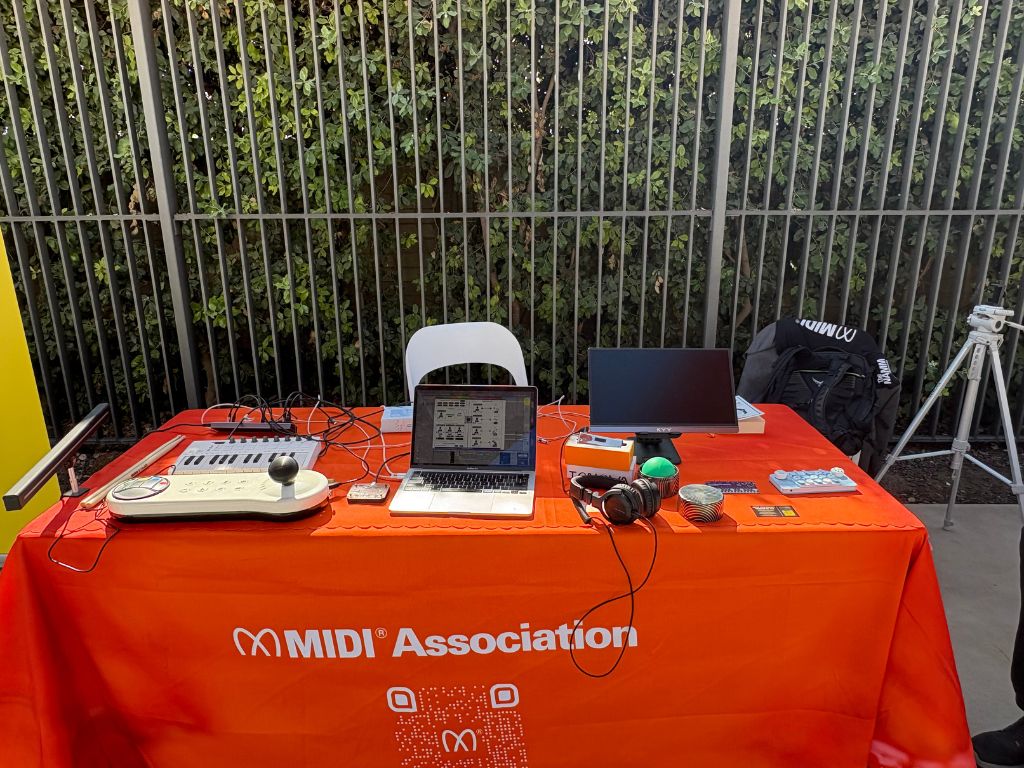 A table with a red MIDI Association tablecloth holds a laptop, small keyboard, monitor, headphones, joystick, cards, and electronics. A white chair and metal fence with greenery are in the background.