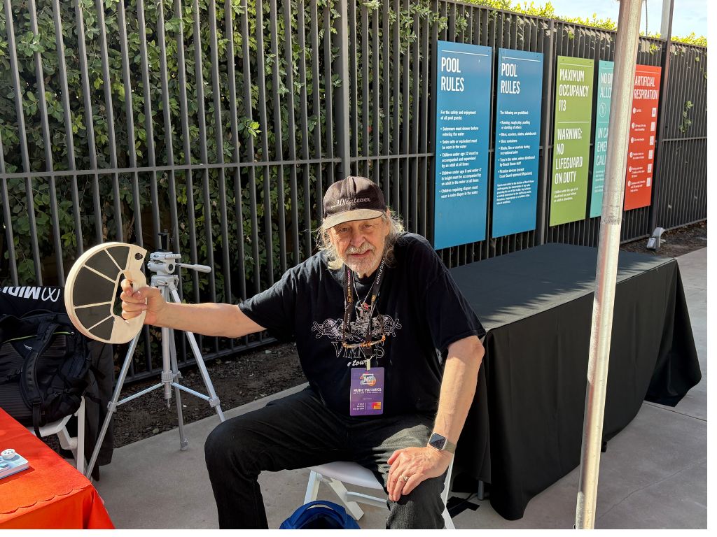 An older man with long gray hair and a beard sits on a chair outdoors, holding a white circular object. He wears a black hat, black shirt, and a lanyard. Colorful pool rule signs and a black table are visible behind him.