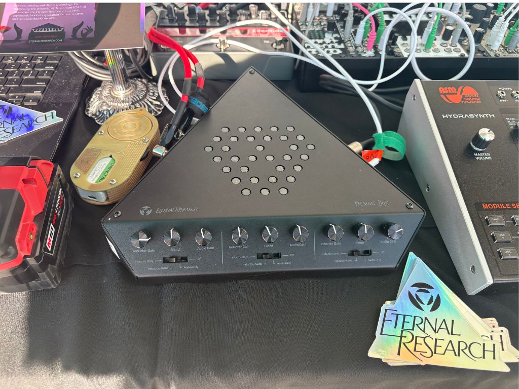 A triangular black audio device with various knobs and vents sits on a table among synthesizers, cables, stickers, and electronic equipment, including an Eternal Research sticker in the bottom right corner.