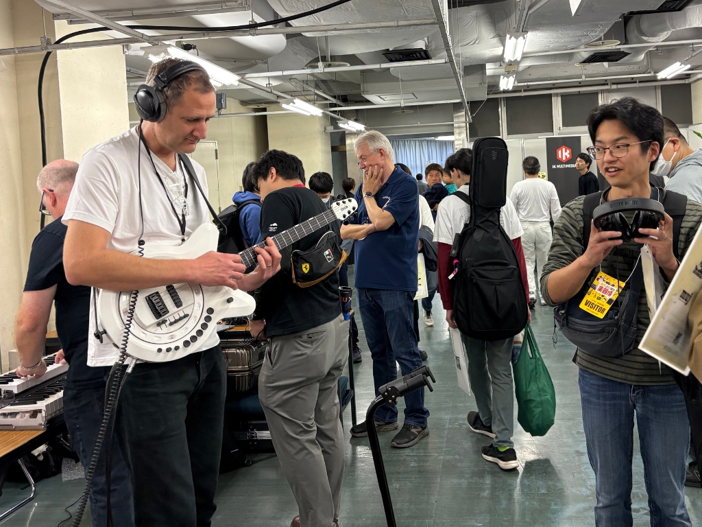 A man wearing headphones plays an electric guitar at an indoor event while others watch, talk, and interact with musical equipment in a busy, well-lit room.