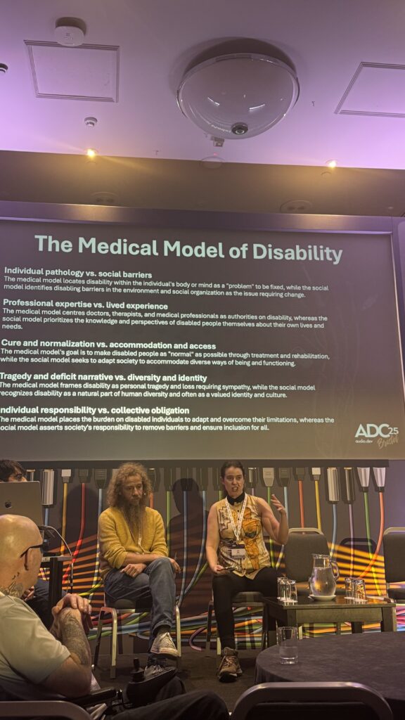 A panel discussion on the Medical Model of Disability. Two speakers sit on stage, with a presentation slide behind them listing key points about disability models. Audience members are visible in the foreground.