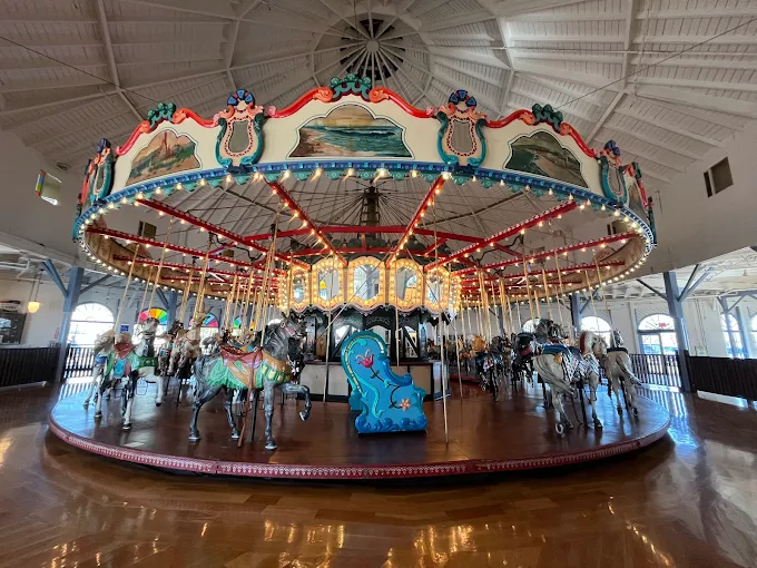 A brightly lit indoor carousel with colorful, ornate horses and a blue sea creature seat, set on a polished wooden floor under a white, domed ceiling with visible beams.