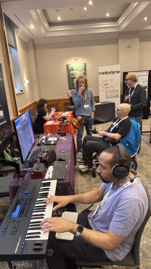 A man plays a keyboard synthesizer at a music technology booth, while others watch and interact. Computers, audio equipment, and banners are visible in a busy conference room.