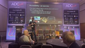 Jay Pocknell from SoundWithoutSight presents on stage at the ADC conference, discussing accessibility. A slide behind them references a TEDx Talk and features images of a vintage phone and retro equipment. Audience members watch and listen.