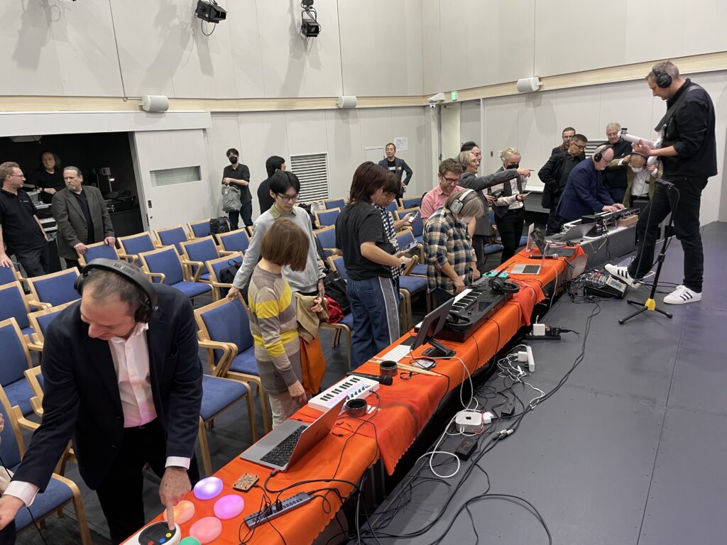 A group of people stands and interacts with electronic musical instruments and equipment on tables covered with orange cloths in a room with chairs and stage lighting. Some people are wearing headphones and appear focused.