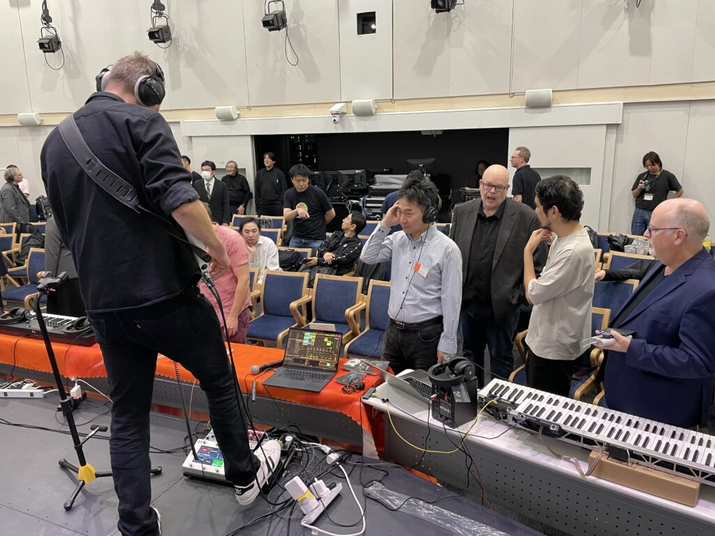 A musician with headphones stands on stage facing sound equipment, while several people, some standing and some seated, observe and interact around a table with electronic gear and keyboards in a studio or auditorium setting.
