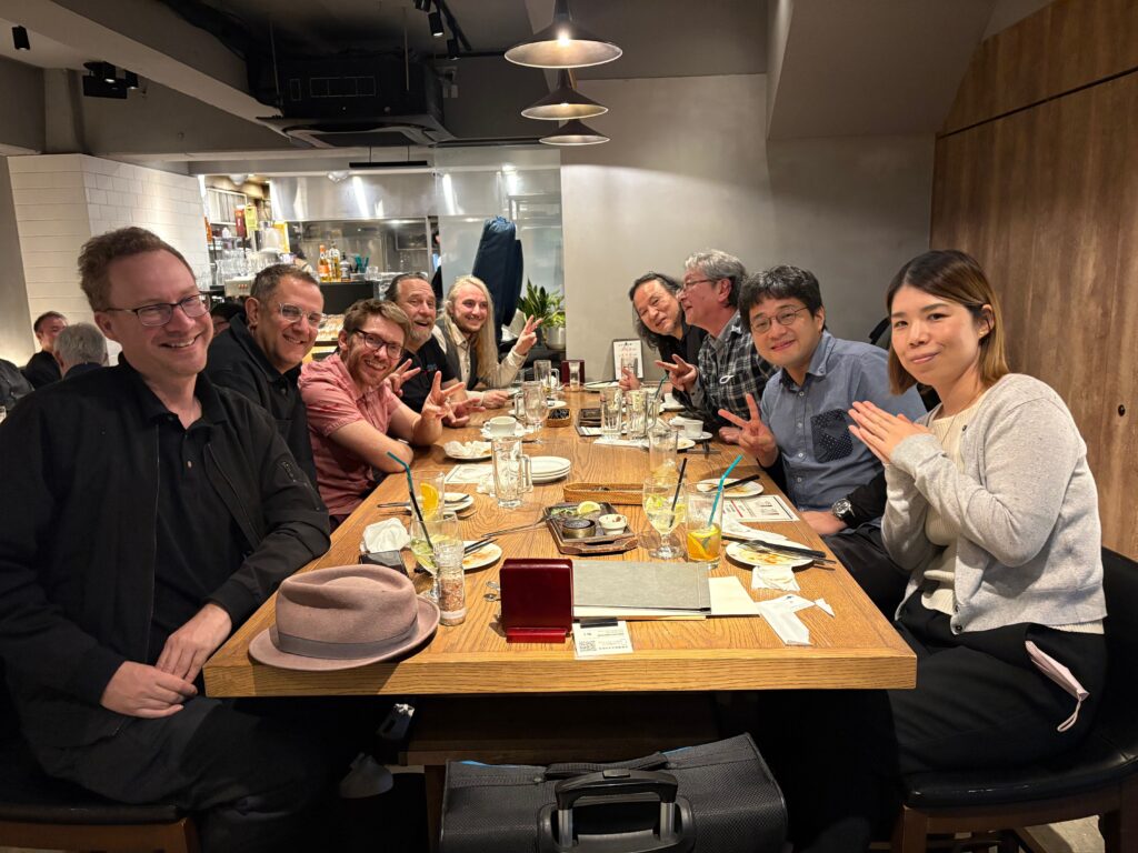 A group of eleven people sit around a restaurant table, smiling and posing for a photo. Plates, drinks, and menus are on the wooden table. The setting is casual with warm lighting and a modern interior.