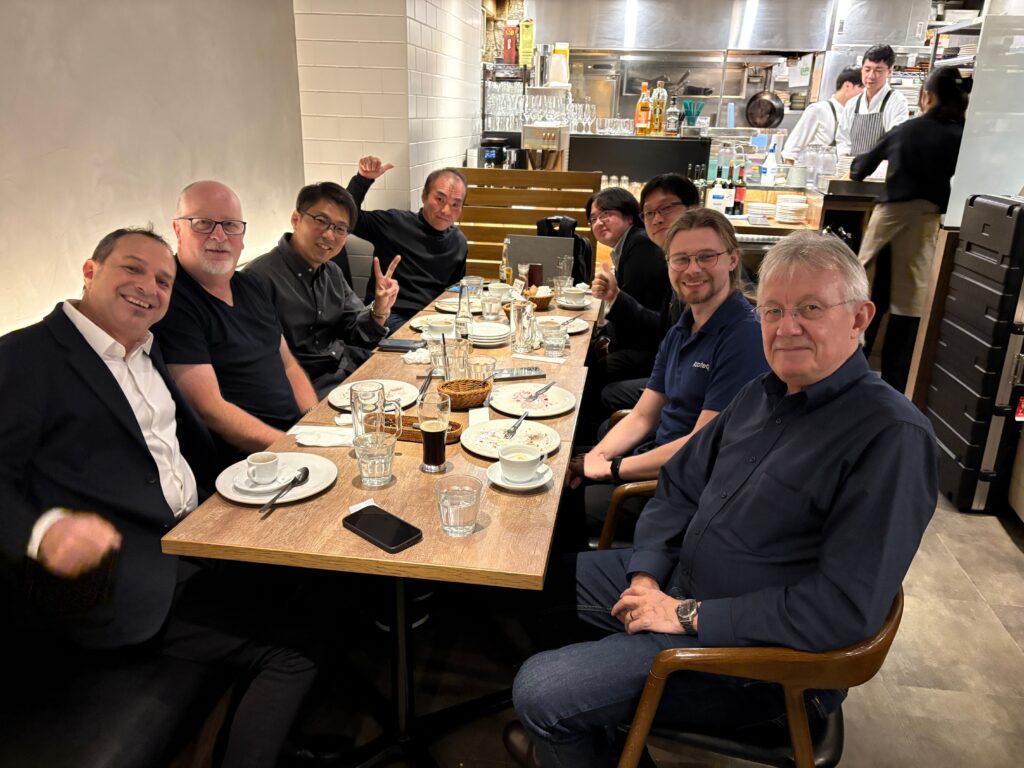 A group of eight men are sitting around a restaurant table with empty plates and drinks, smiling and posing for a photo. The setting is casual, with an open kitchen and restaurant staff visible in the background.