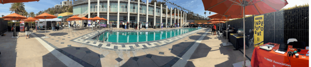 A panoramic view of an outdoor pool area with vendor booths, orange umbrellas, and people walking around. A modern building and palm trees are visible in the background under a clear blue sky.
