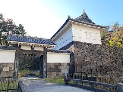 A traditional Japanese castle gate and watchtower with white walls, gray tiled roofs, and stone foundations, surrounded by trees and under a clear blue sky.