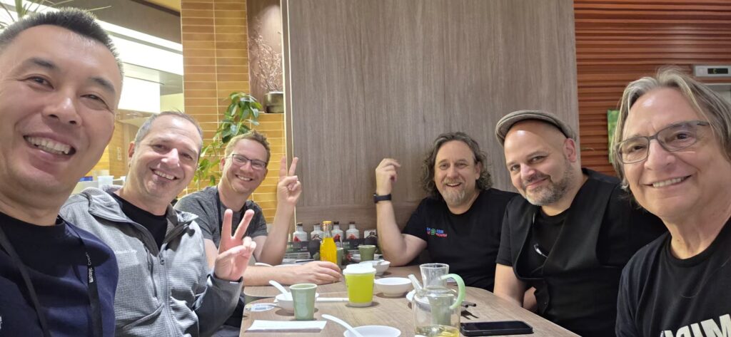 Six smiling men sit around a restaurant table with drinks and dishes, posing for a group selfie. Some are making peace signs, and the atmosphere is casual and friendly.