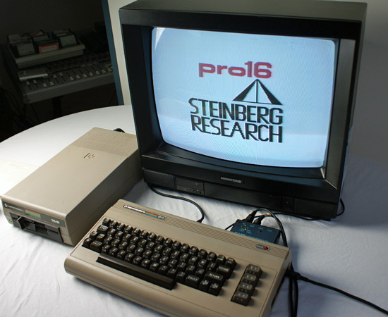 A vintage computer setup with a Commodore 64, a floppy disk drive, and a CRT monitor displaying the “pro16 Steinberg Research” logo. The devices are arranged on a white surface.