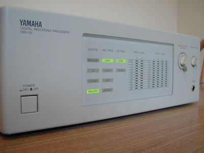 A close-up of a Yamaha digital recording processor, model DCR-D6, with a display panel showing various input options and indicators. The power switch is on and the unit is placed on a wooden surface.