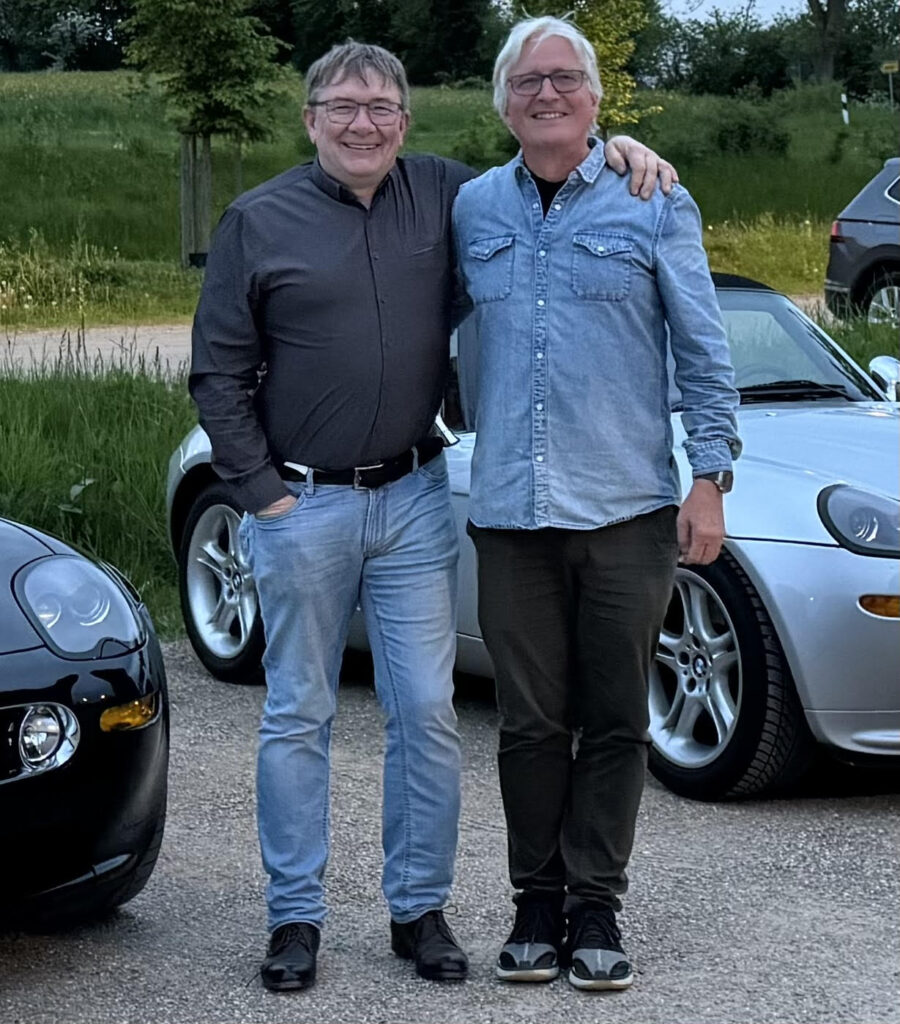 Two smiling men stand close together outdoors, one with his arm around the other. They are in front of two parked sports cars on a paved area, with greenery and trees in the background.