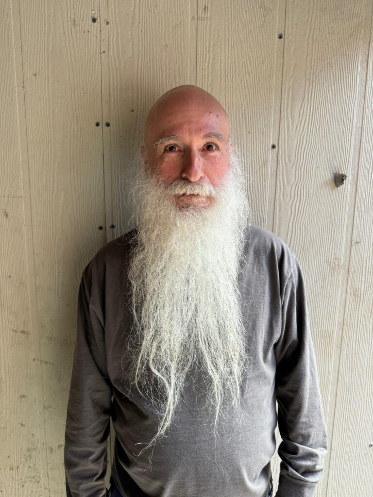An older man with a long white beard and bald head stands against a textured, light-colored wooden wall, wearing a gray long-sleeve shirt and looking at the camera with a slight smile.