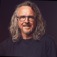 A middle-aged man with long, wavy gray hair and glasses smiles at the camera. He is wearing a black t-shirt and is set against a dark, neutral background.