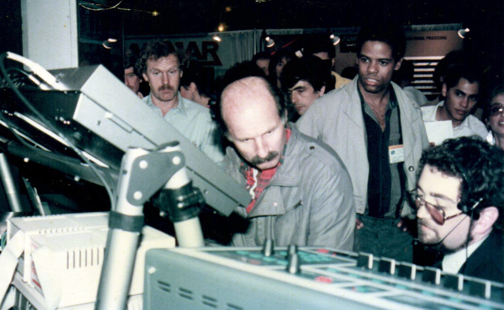 A group of men closely observe electronic music equipment at a trade show or expo, with one man in sunglasses seated and others standing around him, focused on the devices.