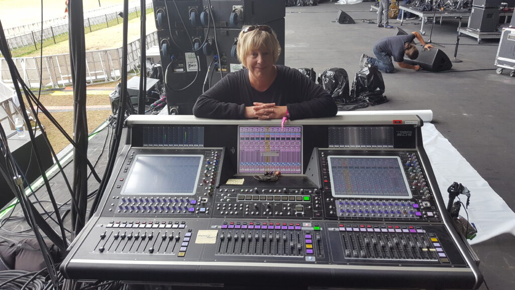 A person with short blonde hair stands behind a large audio mixing console on an outdoor concert stage, with screens and colored buttons visible, while another person works in the background.