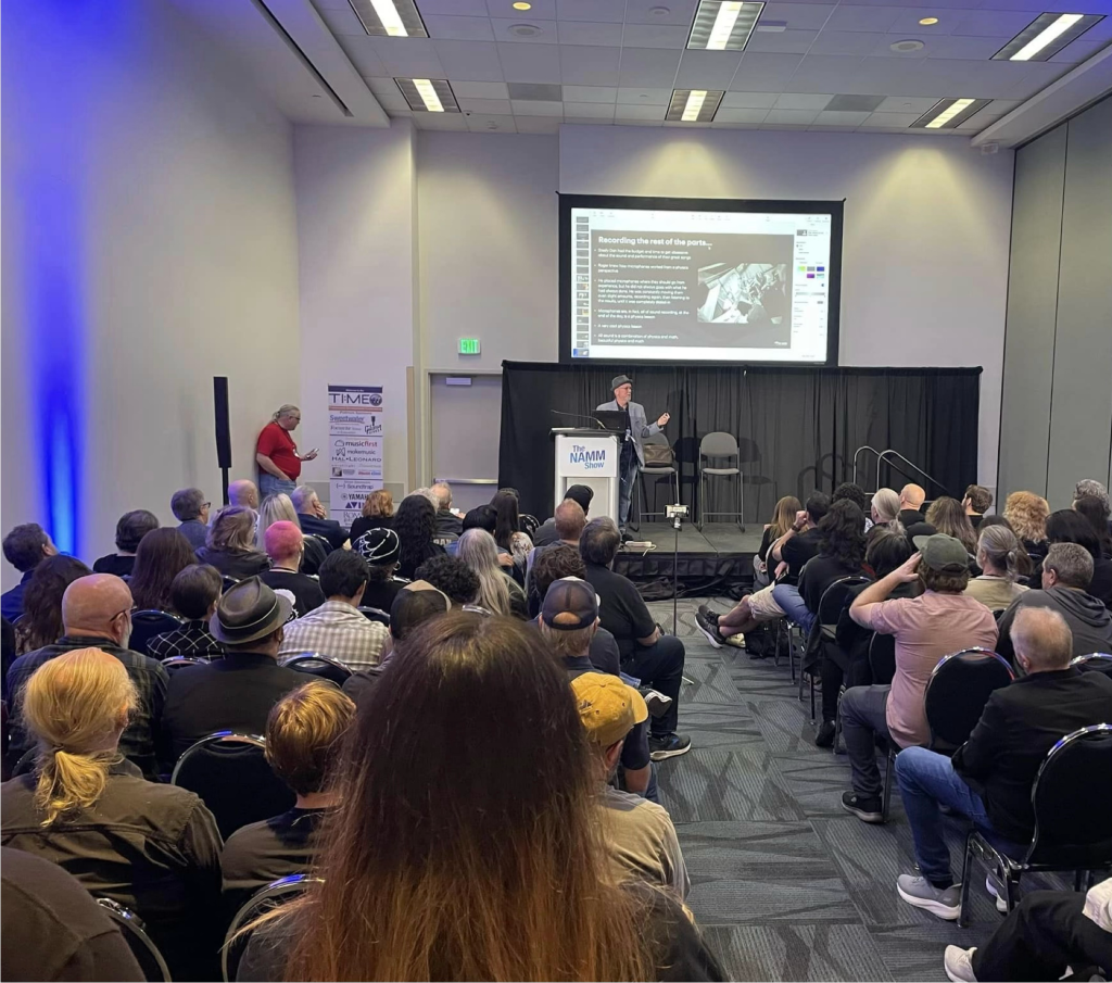 A speaker presents to a seated audience in a conference room, pointing at a projected slide. The crowd faces the stage, and the room is brightly lit with rows of ceiling lights.