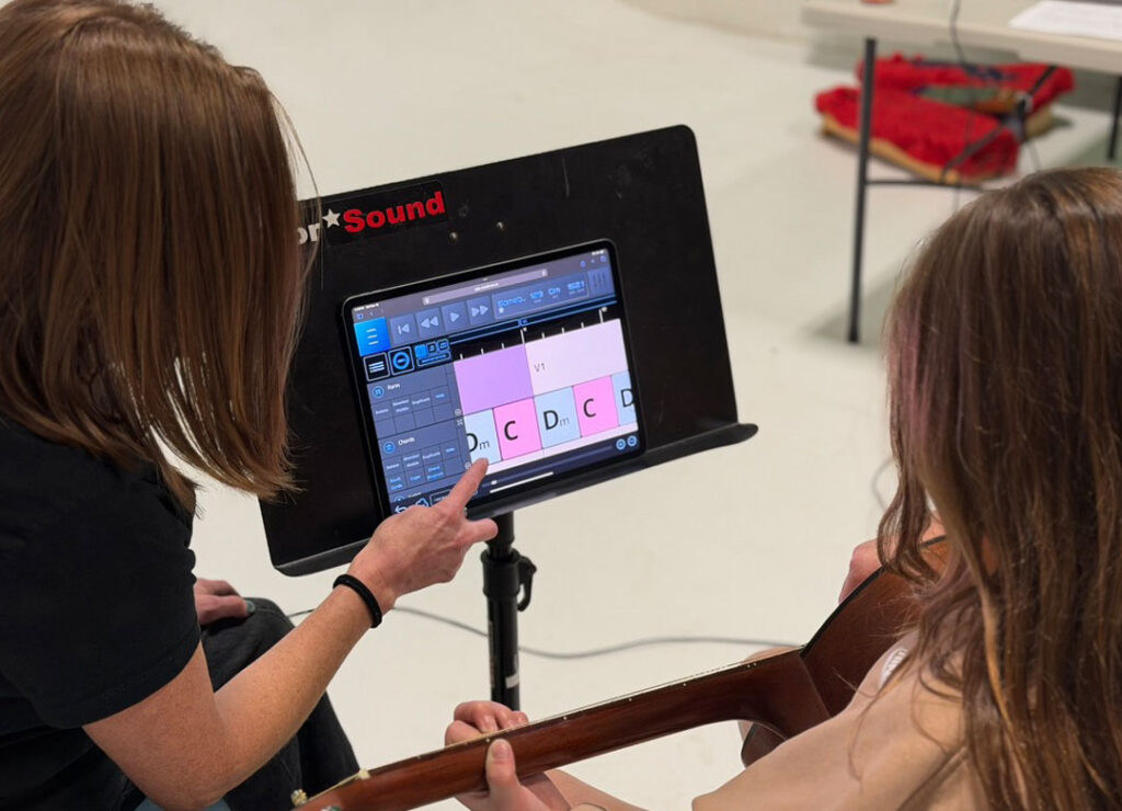A woman points to guitar chords displayed on a tablet screen mounted on a stand, while another person holding a guitar looks on, suggesting a music lesson or practice session.