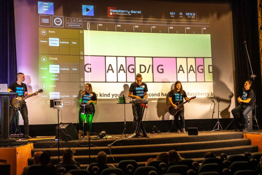 Six musicians perform on stage in front of a large screen showing digital music notation for Raspberry Beret. The audience watches from their seats in a dimly lit theater.