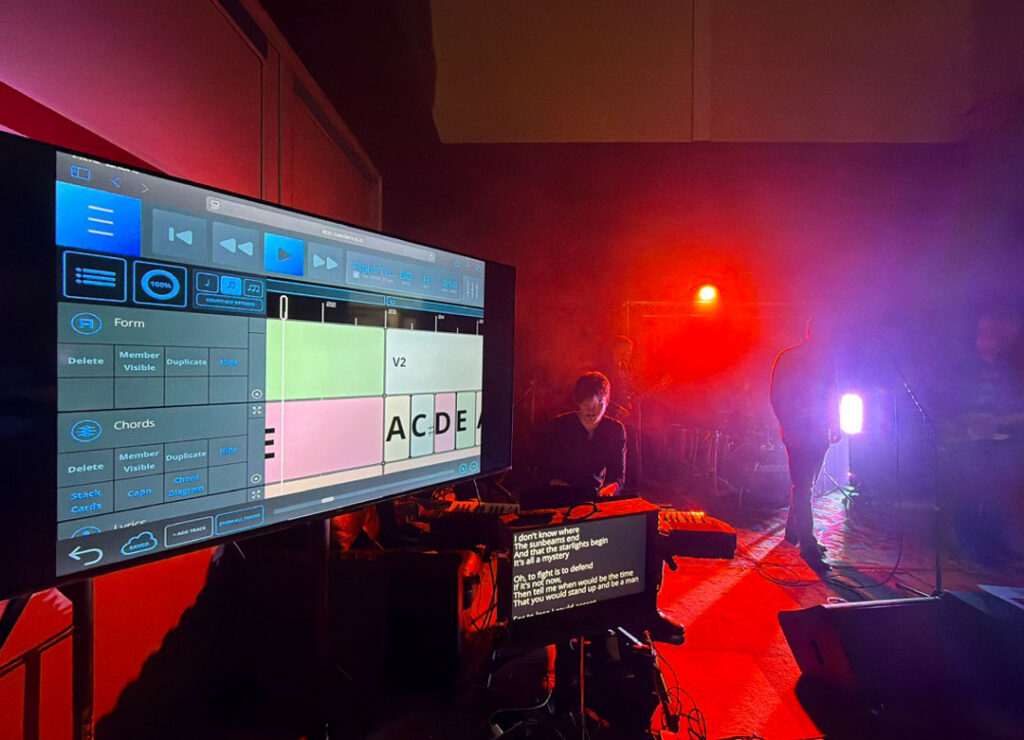 A dimly lit stage with red lighting features a large screen displaying music software, a musician seated nearby, and a monitor showing song lyrics. Musical equipment and cables are visible in the background.