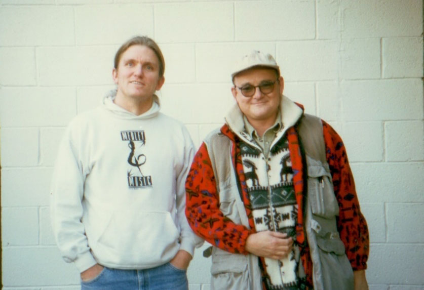 Two men stand in front of a white brick wall. One wears a white hoodie and jeans, the other wears glasses, a light cap, and a patterned jacket with a vest. Both are smiling and appear relaxed.