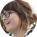 A person with shoulder-length brown hair and glasses is smiling and looking up. They are wearing a white and black patterned top. The background is blurred.