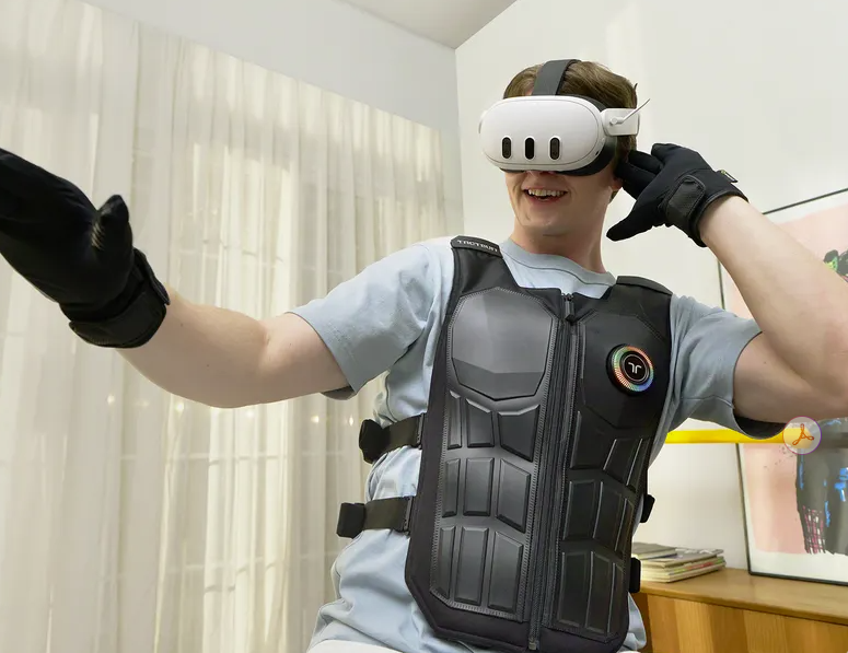 A person wearing a virtual reality headset, haptic vest, and gloves stands indoors, smiling and extending one arm as if interacting with a virtual environment.