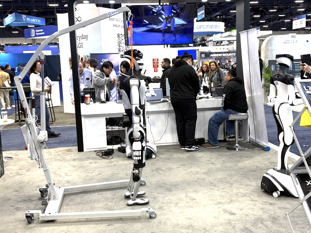 A robotics expo booth displays a robotic exoskeleton on a frame, with several people interacting nearby. A large screen shows tech visuals above the booth, and more booths and visitors are visible in the background.