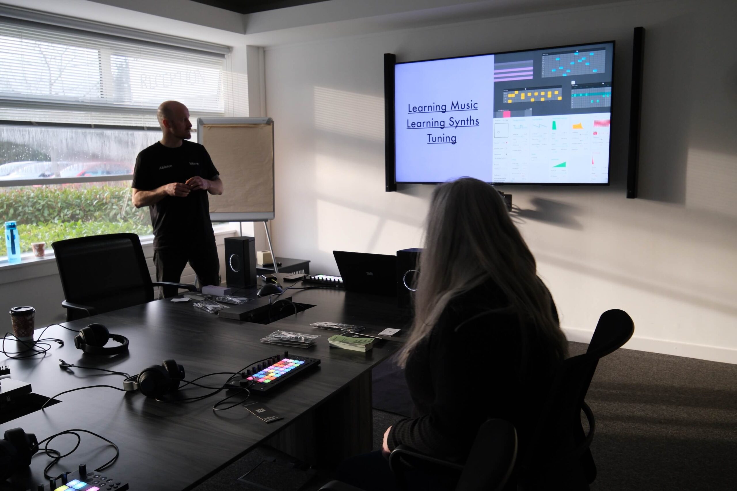 A person stands near a flip chart leading a presentation on Learning Music, Learning Synths, Tuning to an attendee seated at a table with music equipment, while slides are displayed on a large screen.