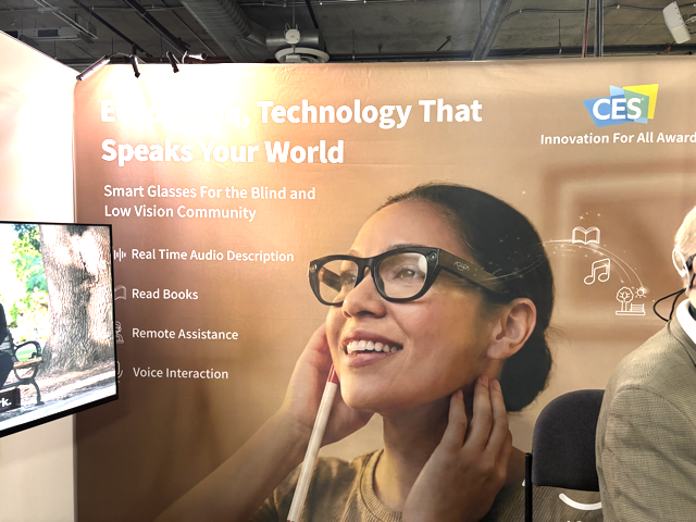 A smiling woman wears smart glasses and touches her ear. The display behind her promotes smart glasses for the blind and low vision community at CES, highlighting features like audio description and voice interaction.