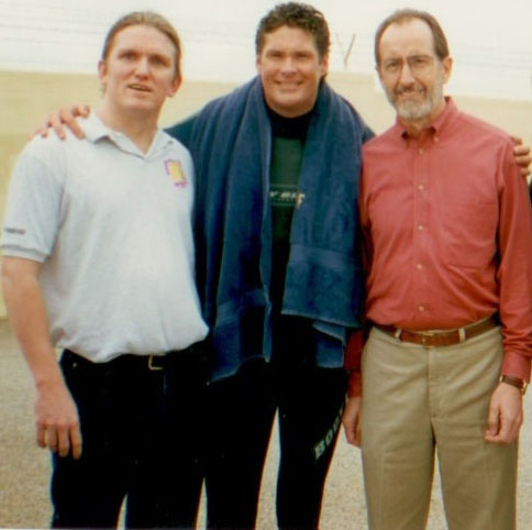 Three men stand outdoors, posing together and smiling. The man in the middle has a blue towel draped over his shoulders. The man on the left wears a white polo shirt, and the man on the right wears glasses and a red button-down shirt.