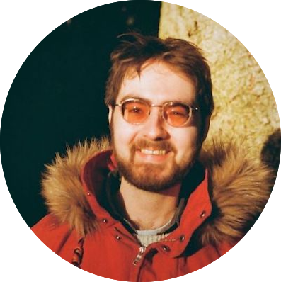 A person with a beard and glasses, wearing a red parka with a fur-lined hood, is smiling at the camera. The background is dark with a light-colored vertical object behind them.
