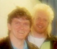 Two men are standing close together and smiling. The photo is blurry and has a soft, hazy focus. One man has dark hair and the other has light blond hair. Both appear to be wearing jackets.