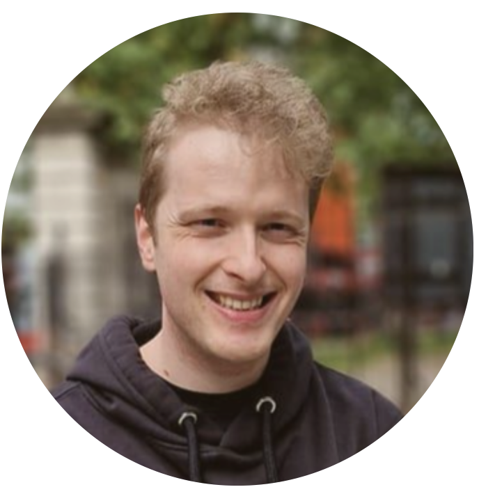 A young person with short, curly blond hair and a black hoodie smiles outdoors, with blurred greenery and buildings in the background.