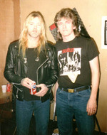 Two men stand side by side indoors. The man on the left has long blond hair, a black leather jacket, and holds a soda can. The man on the right has brown hair and wears a black graphic t-shirt and jeans.
