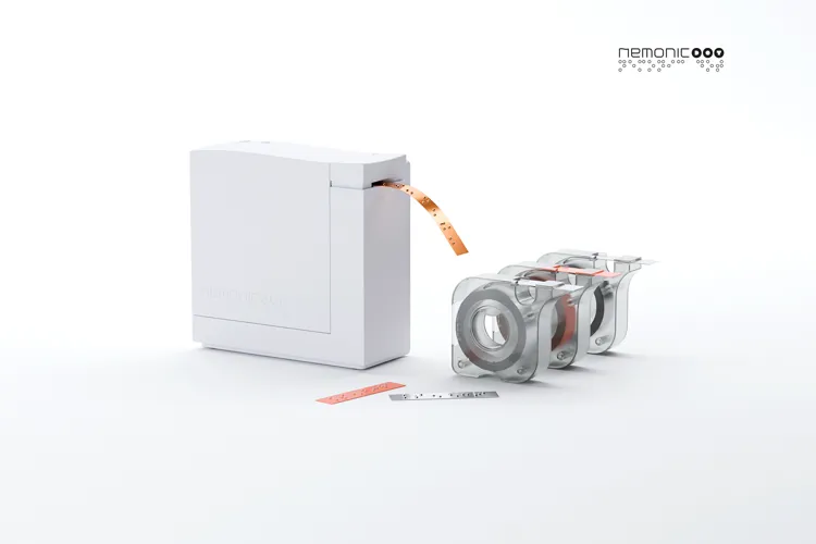 A white compact label printer feeds out an orange printed label. Beside it are three transparent tape rolls on a holder, with one roll partially unwound. The memonic logo appears in the top right corner.