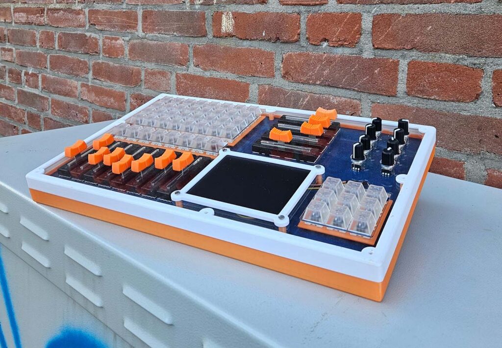 A custom-built electronic music controller with orange and clear buttons, knobs, and sliders sits on a gray surface against a brick wall. The device has a rectangular design with a large central touchscreen.
