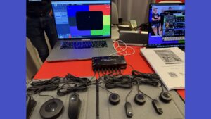 A table with electronic devices, including sensors with wires, a signal processor, an open laptop displaying colorful software, and a monitor showing music software and two people. A booklet and papers are on the right side.