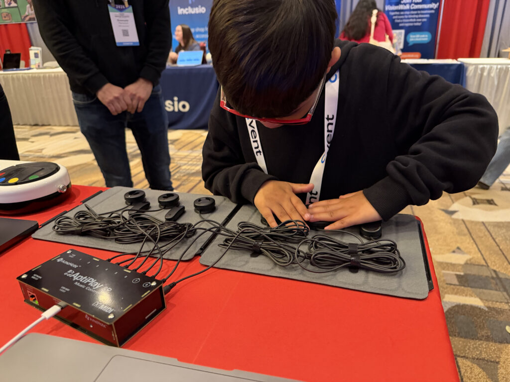 A child wearing glasses uses their hands to interact with a tactile electronic device featuring multiple buttons and wires at an event, with informational booths visible in the background.
