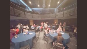A group of people sit and stand around round tables covered in blue tablecloths in a spacious, modern room with high ceilings and a balcony, suggesting a social or networking event.