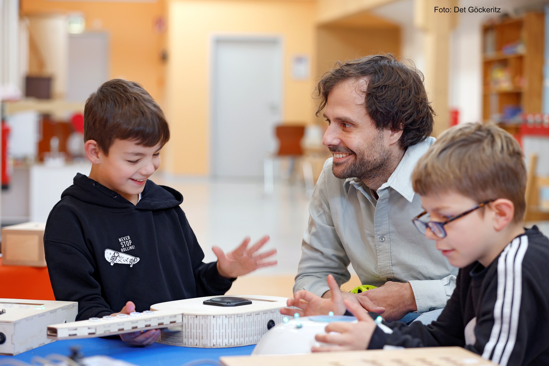 A smiling teacher sits at a table with two young boys, engaging in a hands-on learning activity with electronic devices and kit parts in a bright classroom. One boy gestures animatedly while the other focuses on his project.