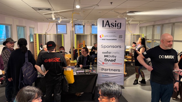A group of people gather around tables and a tall IA-SIG banner with sponsor logos in a brightly lit room at an indoor event. Some attendees converse while others browse materials on the tables.